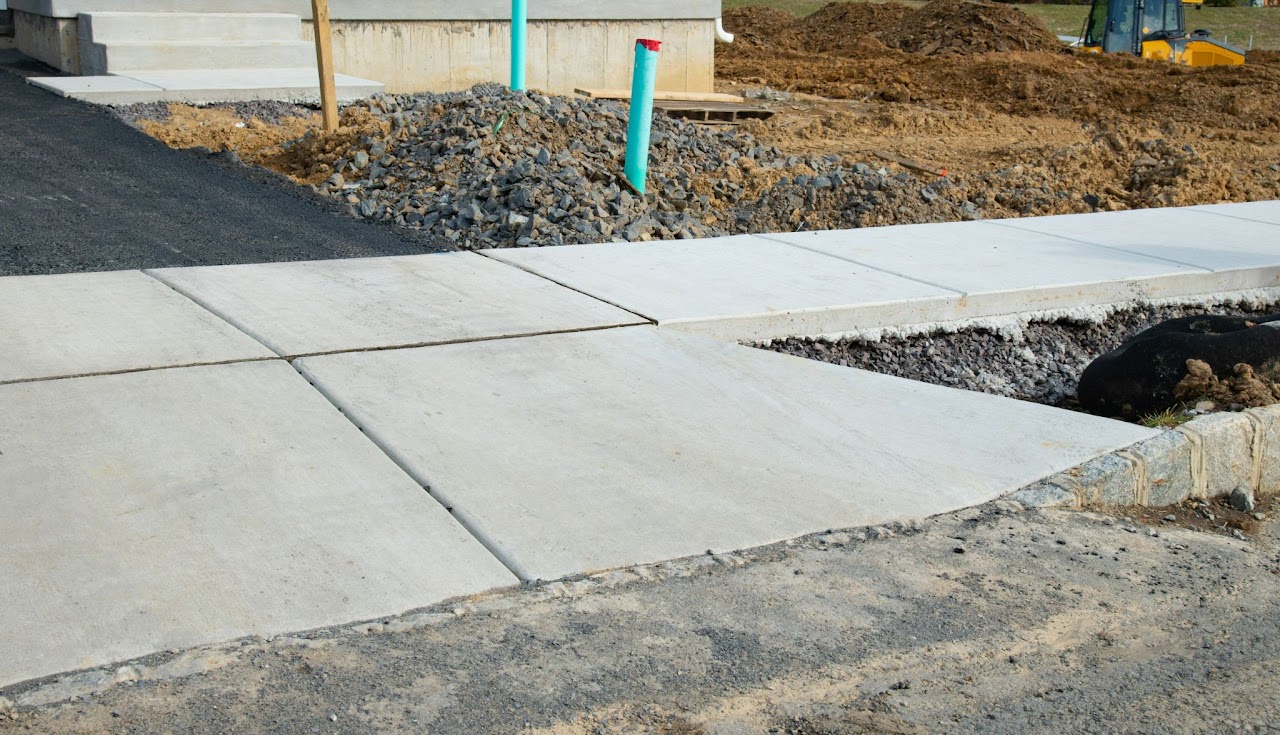 A freshly installed concrete sidewalk and curb under construction, with gravel and soil in the background, showcasing ongoing work by Morris Town Paving's professionals to highlight sidewalk and curb installation and repair services in Morris County, NJ.