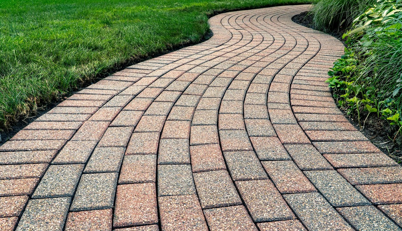 A winding paver walkway installed by Morristown Paving in Morris County, NJ. The curved path features tan-colored pavers in an elegant curved pattern with dark borders, bordered by lush green lawn and landscaping