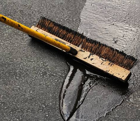 Close-up of a professional sealcoating brush applying dark protective sealant to asphalt by Morristown Paving in Morris County, NJ. The image shows the wet application process, demonstrating the company's pavement maintenance and protection services.