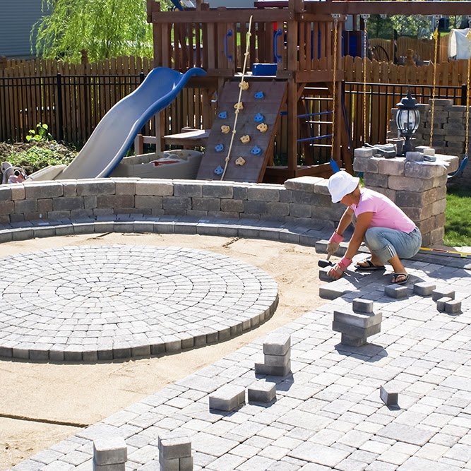 A Morristown Paving contractor in Morris County, NJ installs a circular paver patio with a curved retaining wall near a backyard playground. The pavers are being laid in a decorative circular pattern, showcasing the precise work in progress.