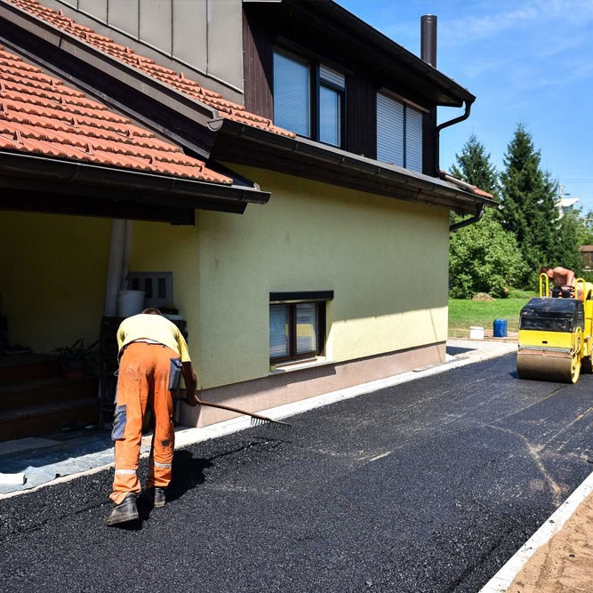 A Morristown Paving crew member, performing services for a client in Morris County, NJ, uses a rake tool to spread fresh asphalt on a residential driveway. The company's paving machine and the client home are visible in the background as the worker ensures even asphalt distribution.