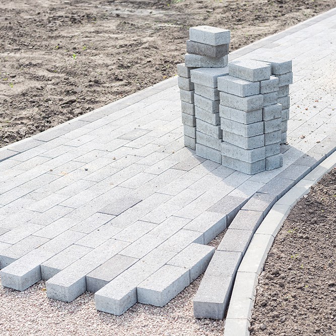 A Morristown Paving project in Morris County, NJ shows the installation process of a paver walkway. Gray concrete pavers are partially laid in a neat pattern, with stacked extra pavers and exposed dirt visible, demonstrating the professional work in progress.