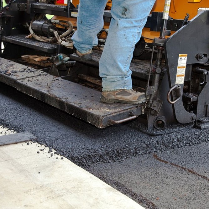 Close-up view of a Morristown Paving crew member's work boots standing on the screed plate of an asphalt paving machine at a residence in Morris County, NJ. Fresh black asphalt is being laid onto a driveway surface, showing the precision of the paving process.